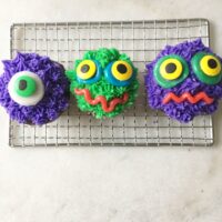 a trio of cupcakes decorated like monsters in shades of purple and green