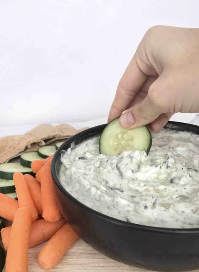 hand dipping cucumber into a bowl of tzatziki 