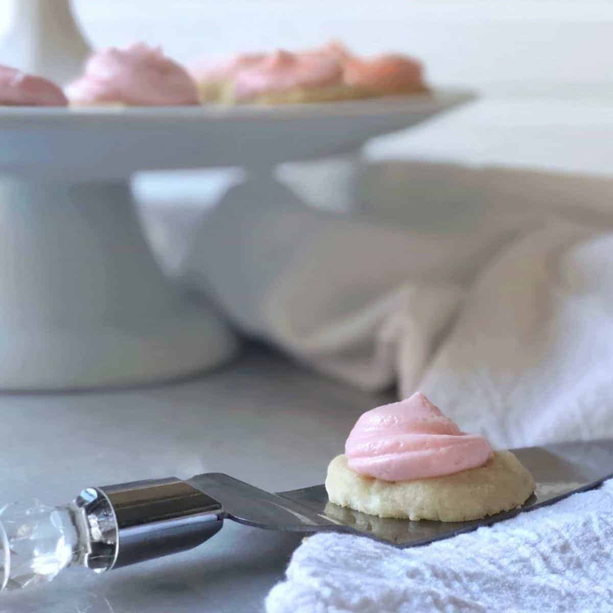 tiny sugar sugar cookie with swirl of pink frosting sitting on cake server with background platter of more cookies.