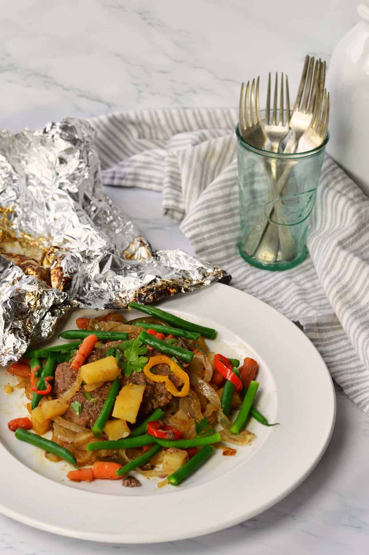 white plate with fully cooked foil dinner and piece of scrunched aluminum foil. 