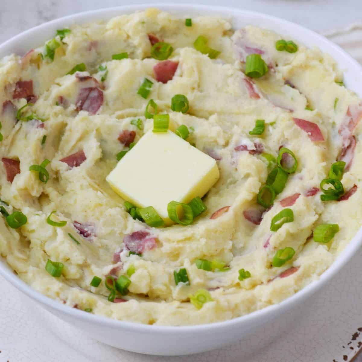 White bowl filled with a swirl of creamy roasted garlic red skin mashed potatoes garnished with green onion and a square of butter.